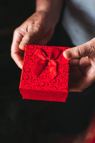 Close-up of elderly female hands holding red gift box. Black, dark background