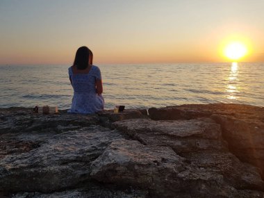 Beyaz elbiseli bir kız sahilde bir taşın üzerinde oturuyor ve gün batımını izliyor. Sarı-turuncu güneş ufukta asılı