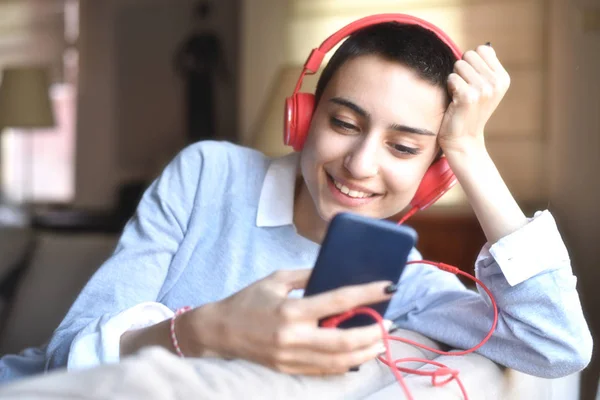 Müzik dinleyen genç bir kadın. Kulağında kırmızı kulaklıkla gülümsüyor. Elinde telefonla.