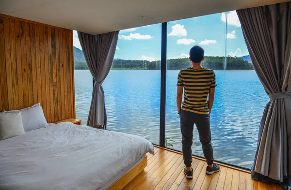 An Asian young man relaxing at wooden resort near lake with mountain background.