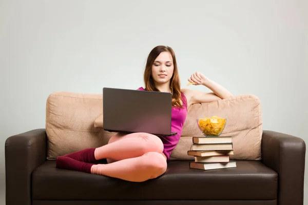 ピンクの服を着た若い女性は 自宅のソファに座ってノートパソコンで作業し 本の上に置かれたポテトチップスを食べていました 灰色の背景を削除しやすい — ストック写真