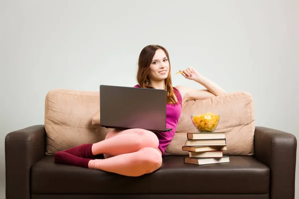 ピンクの服を着た若い女性は 自宅のソファに座ってノートパソコンで作業し 本の上に置かれたポテトチップスを食べていました 灰色の背景を削除しやすい — ストック写真