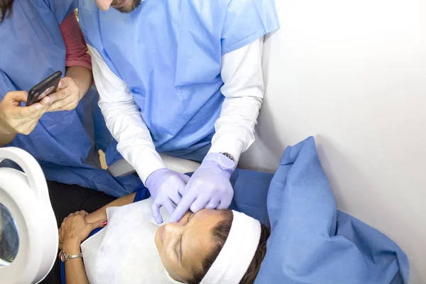 Bir doktor muayenehanesinde bir doktor, bir kadın hastaya estetik tedaviyi mobil leriyle kaydeden birkaç öğrenciye açıklar.