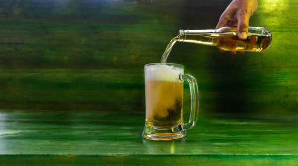 A hand serves beer foaming in a jug with a green textured wooden background