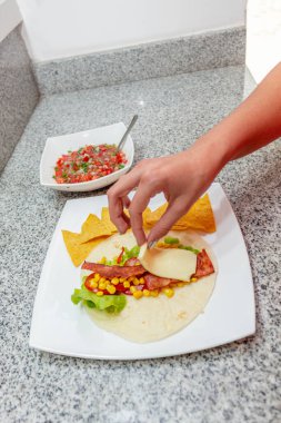 Bir el peyniri domuz pastırmalı ve mısır taneli taconun üzerine koyar. Yanında da pico de gallo olan bir kase.