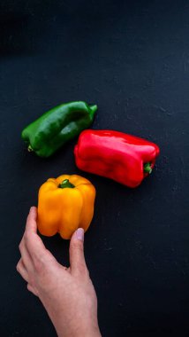 Bir kadın eli, kırsal bir yüzeyde başka bir rengin yanında duran sarı bir biberi tutuyor.