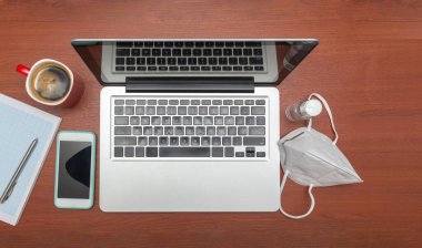 Bilgisayar maske ve dezenfektan jel ile bir iş istasyonunda defter, cep telefonu ve kahve fincanının yanında.