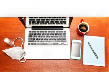 Bilgisayarlı bir masanın üst görüntüsü, cerrahi maske, el dezenfektanı jeli, cep telefonu, evdeki bir ofiste defter.