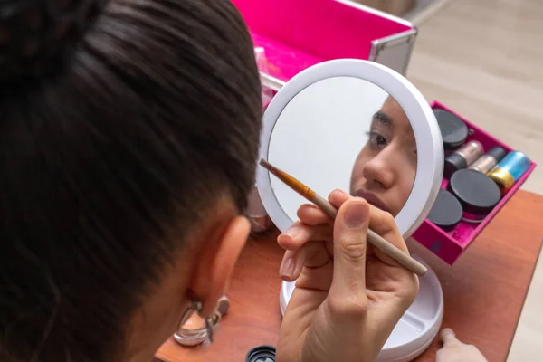 Teenage girl looking through a small mirror while putting on makeup in her living room