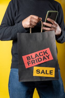 Shopper using smartphone while holding black friday shopping bag against yellow background