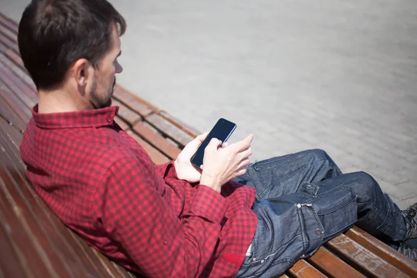Bearded man sitting on bench in park with phone in hands