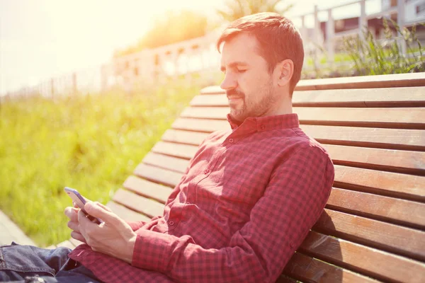 Bearded man sitting on bench in park with phone in hands