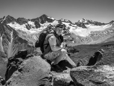 Dağların yükseklerinde bir kayanın üzerinde oturan kameralı bir adam fotoğraf çekiyor..