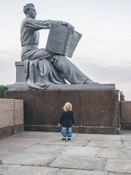 A little girl stands in front of a Soviet monument depicting a man reading an open book.