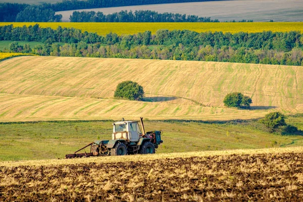 Un tractor agrícola arada un campo con un arado después del trigo cosechado. En el fondo hay campos de girasoles . —  Fotos de Stock