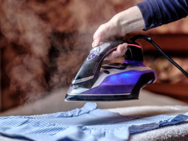 Kadın eli, masadaki kıyafetleri ütülüyor. Ütüden çıkan sıcak buhar. Koyu bulanık arkaplan.