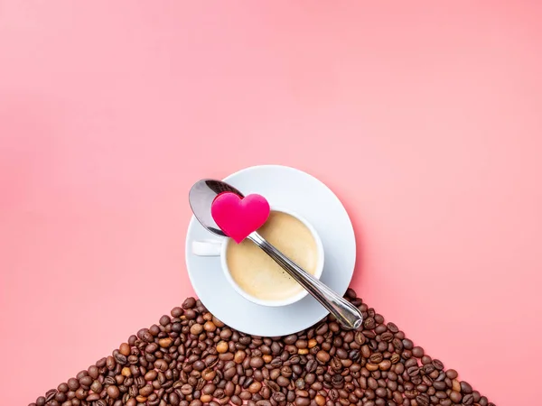 Flat lay. A white ceramic cup with coffee stands on a hill of coffee beans on a pink background. On a cup lies a metal spoon and a small silk heart. Fragrant roasted coffee for breakfast. Copy space.