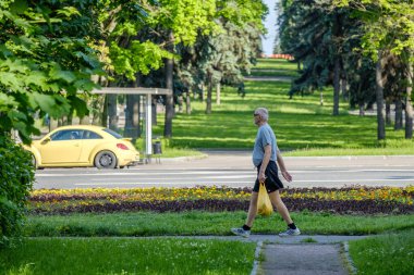 Moskova, Rusya. 14 Haziran 2020. Elinde sarı bir çanta olan tişörtlü ve şortlu bir adam yol boyunca kaldırım boyunca yürüyor. Arka planda bir park ve sarı bir Volkswagen böcek arabası var..