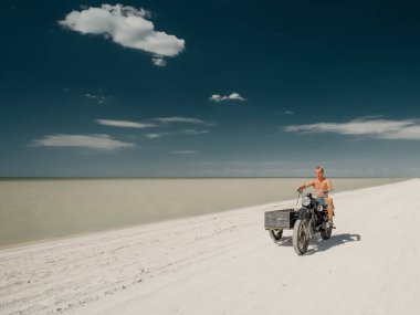 Glafirovka, Krasnodar Bölgesi, Rusya. 29 Temmuz 2020 'de genç bir adam deniz kıyısında beyaz kumlu bir plajda eski bir motosiklete biniyor. Parlak güneş ışığı. Sıcak yaz günü. Derin mavi gökyüzü.