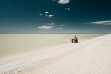 Glafirovka, Krasnodar Bölgesi, Rusya. 29 Temmuz 2020 'de genç bir adam deniz kıyısında beyaz kumlu bir plajda eski bir motosiklete biniyor. Parlak güneş ışığı. Sıcak yaz günü. Derin mavi gökyüzü.