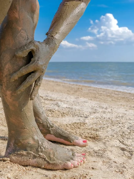 Селективный акцент на красивой женской ноге с ярко-красным педикюром на пляже песка. Руки мажут заживляющую грязь на лодыжках ног. Омоложение кожи во время отдыха в море. Спа. копировальное пространство — стоковое фото