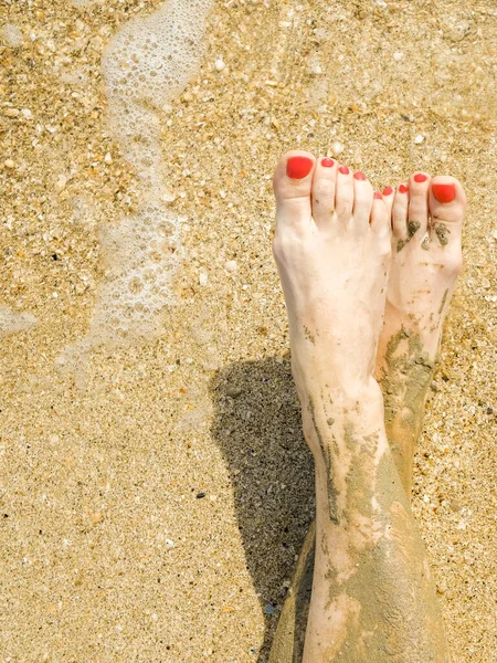Vista dall'alto di bei piedi femminili con pedicure rosso brillante sulla sabbia della spiaggia. Le caviglie dei piedi sono imbrattate di fango medicinale. Ringiovanimento della pelle durante il relax in mare. Spa. Copia spazio — Foto Stock