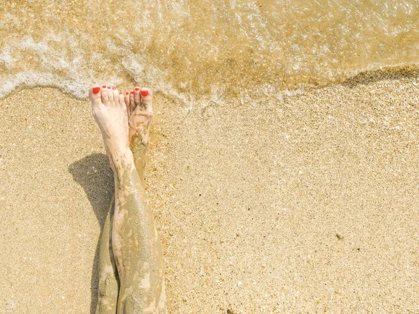 Vista dall'alto di bei piedi femminili con pedicure rosso brillante sulla sabbia della spiaggia. Le caviglie dei piedi sono imbrattate di fango medicinale. Ringiovanimento della pelle durante il relax in mare. Spa. Copia spazio — Foto Stock