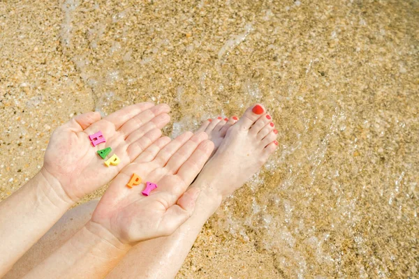 Donna mani e piedi con una pedicure rossa sullo sfondo di sabbia marina e surf. Nei palmi delle lettere con l'iscrizione Happy. Vacanze di vacanza. Copia spazio. — Foto Stock