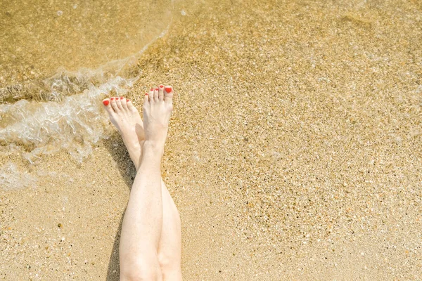 Vista dall'alto di bei piedi femminili con pedicure rosso brillante sulla sabbia della spiaggia. L'onda del mare lava i piedi alle donne. Relax e divertimento durante la vostra vacanza al mare. Copia spazio. — Foto Stock