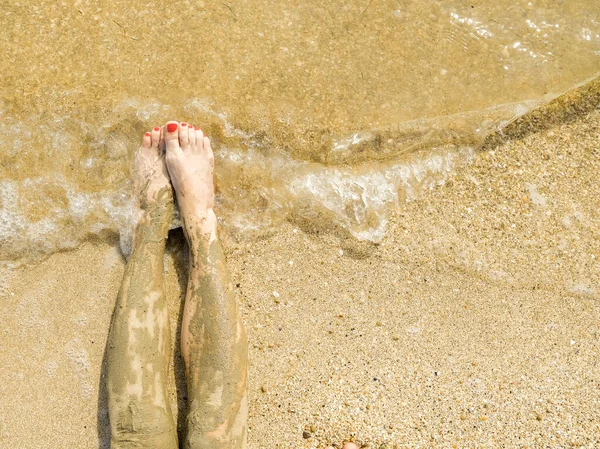 Vista superior de hermosos pies femeninos con pedicura de color rojo brillante en la arena de la playa. Los tobillos de los pies están untados con barro medicinal. Rejuvenecimiento de la piel mientras se relaja en el mar. Spa. Copiar espacio — Foto de Stock