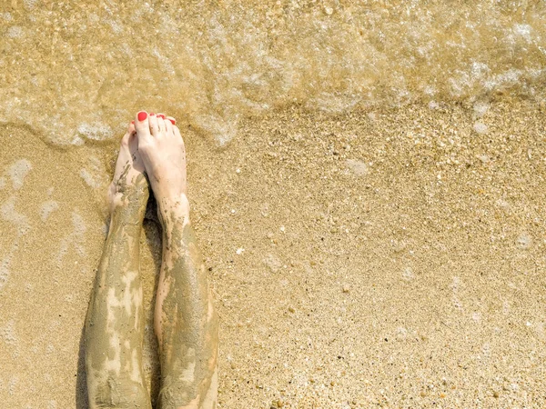 Vista dall'alto di bei piedi femminili con pedicure rosso brillante sulla sabbia della spiaggia. Le caviglie dei piedi sono imbrattate di fango medicinale. Ringiovanimento della pelle durante il relax in mare. Spa. Copia spazio — Foto Stock