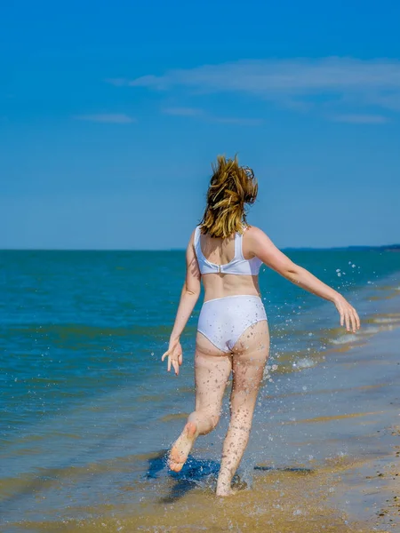 Hátsó kilátás egy lány fut a távolba mentén homokos tengerparton fröccsenő tengervíz. A lány fehér fürdőruhát és napszemüveget visel. Szabadság. Kültéri fitnesz. Nyári napsütéses nap. — Stock Fotó