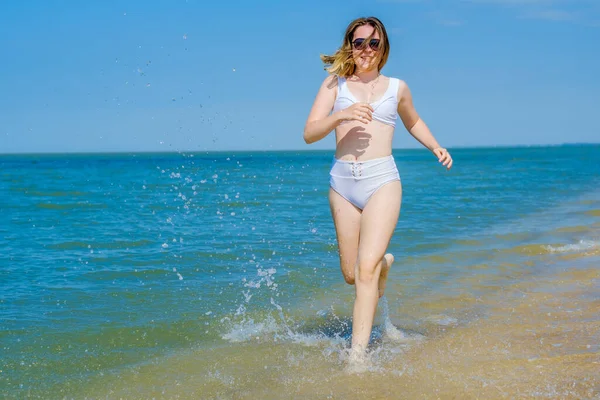 Uma jovem corre ao longo da praia de areia ao longo do surf. Salpicos de água do mar. A menina está usando um maiô branco e óculos de sol. Liberdade e descuido. Aptidão exterior. Verão dia ensolarado — Fotografia de Stock