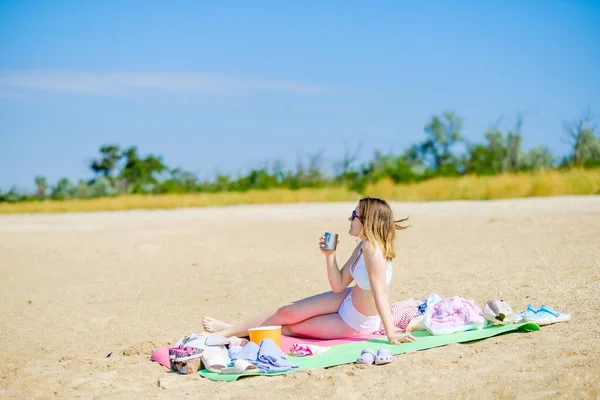 Beyaz mayo giyen seksi genç kız elinde kupayla bir paspasın üzerinde oturuyor ve içiyor. Sahilde tek başına kız pikniği. Güneşli bir gün ve beyaz kum. Deniz kenarında bir cennet tatili. — Stok fotoğraf