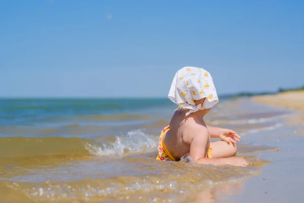 Fokus selektif pada bayi perempuan yang terbaring di air di pasir di pantai. Hari yang cerah. Pandangan ke belakang. Di kepala adalah syal bandana putih. Bidang kedalaman yang dangkal. Masa kecil yang bahagia. Salin ruang — Stok Foto
