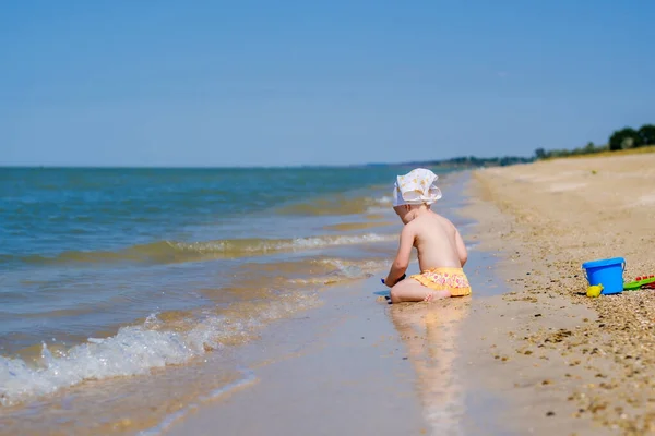 Fokus selektif pada balita gadis duduk di air di pasir di pantai. Anak itu bermain dengan mainan. Pandangan ke belakang. Di kepala adalah syal bandana putih. Bidang kedalaman yang dangkal. Happy childhood — Stok Foto