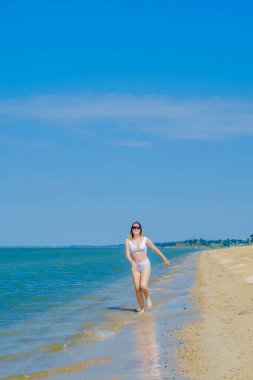 Genç bir kız sörf boyunca kumlu deniz plajı boyunca koşar. Deniz suyu damlaları. Kız beyaz bir mayo ve güneş gözlüğü takıyor. Özgürlük ve dikkatsizlik. Açık hava fitness. Yaz güneşli bir gün