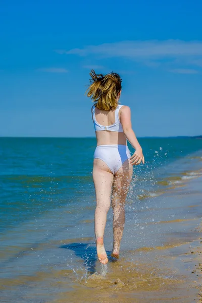 Rückansicht eines Mädchens, das an einem Sandstrand in die Ferne flieht. Das Mädchen trägt einen weißen Badeanzug und eine Sonnenbrille. Freiheit. Fitness im Freien. Sommersonniger Tag. — Stockfoto