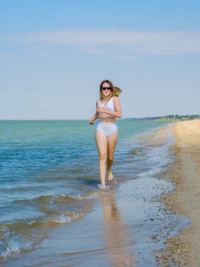 Genç bir kız sörf boyunca kumlu deniz plajı boyunca koşar. Deniz suyu damlaları. Kız beyaz bir mayo ve güneş gözlüğü takıyor. Özgürlük ve dikkatsizlik. Açık hava fitness. Yaz güneşli bir gün