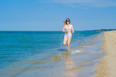 Genç bir kız sörf boyunca kumlu deniz plajı boyunca koşar. Deniz suyu damlaları. Kız beyaz bir mayo ve güneş gözlüğü takıyor. Özgürlük ve dikkatsizlik. Açık hava fitness. Yaz güneşli bir gün