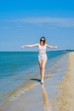 Genç bir kız sörf boyunca kumlu deniz plajı boyunca koşar. Deniz suyu damlaları. Kız beyaz bir mayo ve güneş gözlüğü takıyor. Özgürlük ve dikkatsizlik. Açık hava fitness. Yaz güneşli bir gün