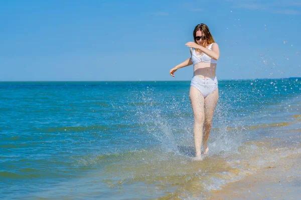 Молодая девушка бежит вдоль песчаного морского пляжа вдоль серфинга. Всплески морской воды. Девушка в белом купальнике и солнечных очках. Свобода и беспечность. Фитнес на открытом воздухе. Солнечный летний день — стоковое фото