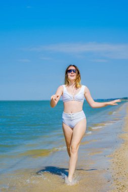 Genç bir kız sörf boyunca kumlu deniz plajı boyunca koşar. Deniz suyu damlaları. Kız beyaz bir mayo ve güneş gözlüğü takıyor. Özgürlük ve dikkatsizlik. Açık hava fitness. Yaz güneşli bir gün