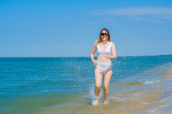 Uma jovem corre ao longo da praia de areia ao longo do surf. Salpicos de água do mar. A menina está usando um maiô branco e óculos de sol. Liberdade e descuido. Aptidão exterior. Verão dia ensolarado — Fotografia de Stock