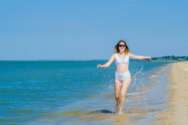 En ung flicka springer längs sandstranden längs surfingen. Vattenstänk i havet. Flickan har en vit baddräkt och solglasögon. Frihet och slarv. Utomhus fitness. Sommaren solig dag — Stockfoto