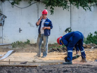 Moskova. - Rusya. 06 Eylül 2020 'de bir inşaat alanında mavi üniformalı ve kırmızı kasklı işçiler öğütücüyle metal kesiyorlar. Yanan metal talaşlarının kıvılcımları uçuşuyor.
