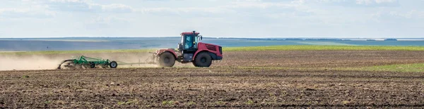 Un tractor rojo de granja en una nube de polvo cultiva el suelo en el campo con un cultivador después de la cosecha. Día soleado de verano. Tierra fértil. Maquinaria agrícola moderna. Copiar espacio. —  Fotos de Stock