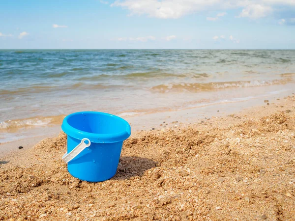 Deniz kenarındaki kumda mavi plastik bebek kovası. Tatil sırasında çocuklarla deniz eğlencesi kavramı. Deniz kıyısındaki tatil köyünde güneşli bir yaz günü. Mavi gökyüzü ve sakin deniz. Boşluğu kopyala