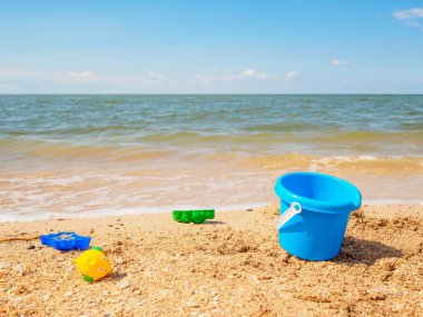 Mavi plastik kova ve deniz kenarındaki kumda bebek oyuncakları. Tatil sırasında çocuklarla deniz eğlencesi kavramı. Deniz kıyısındaki tatil köyünde güneşli bir yaz günü. Mavi gökyüzü ve sakin deniz. Boşluğu kopyala