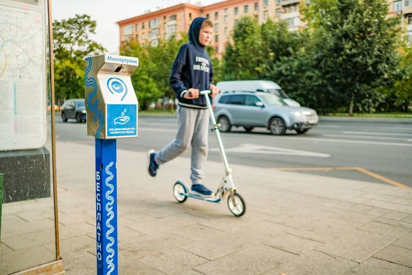 Moskou. Rusland. 04 september 2020 Een apparaat voor het desinfecteren van handen bij een transportstop met een jongen op een scooter en een weg met transport op de achtergrond. Moderne technologieën die mensen beschermen — Stockfoto